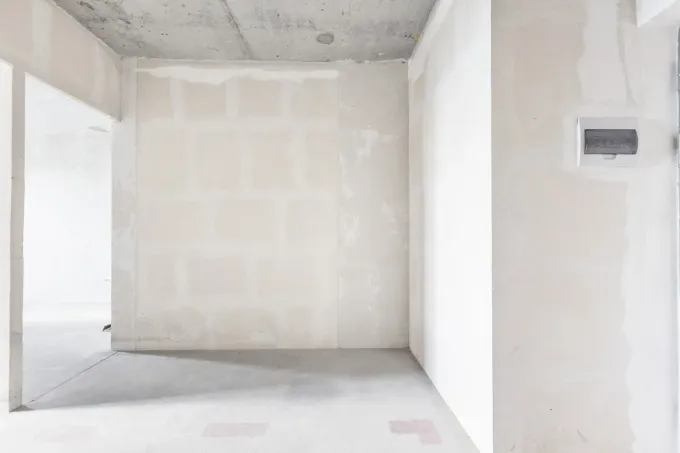 Habitación vacía sin terminar, con paredes de pladur blanco, techo de hormigón y una puerta que se abre a la izquierda.