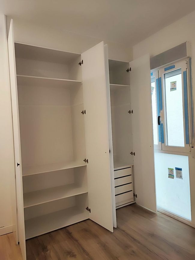 Armario empotrado vacío con las puertas blancas abiertas, en una habitación luminosa junto a una ventana.