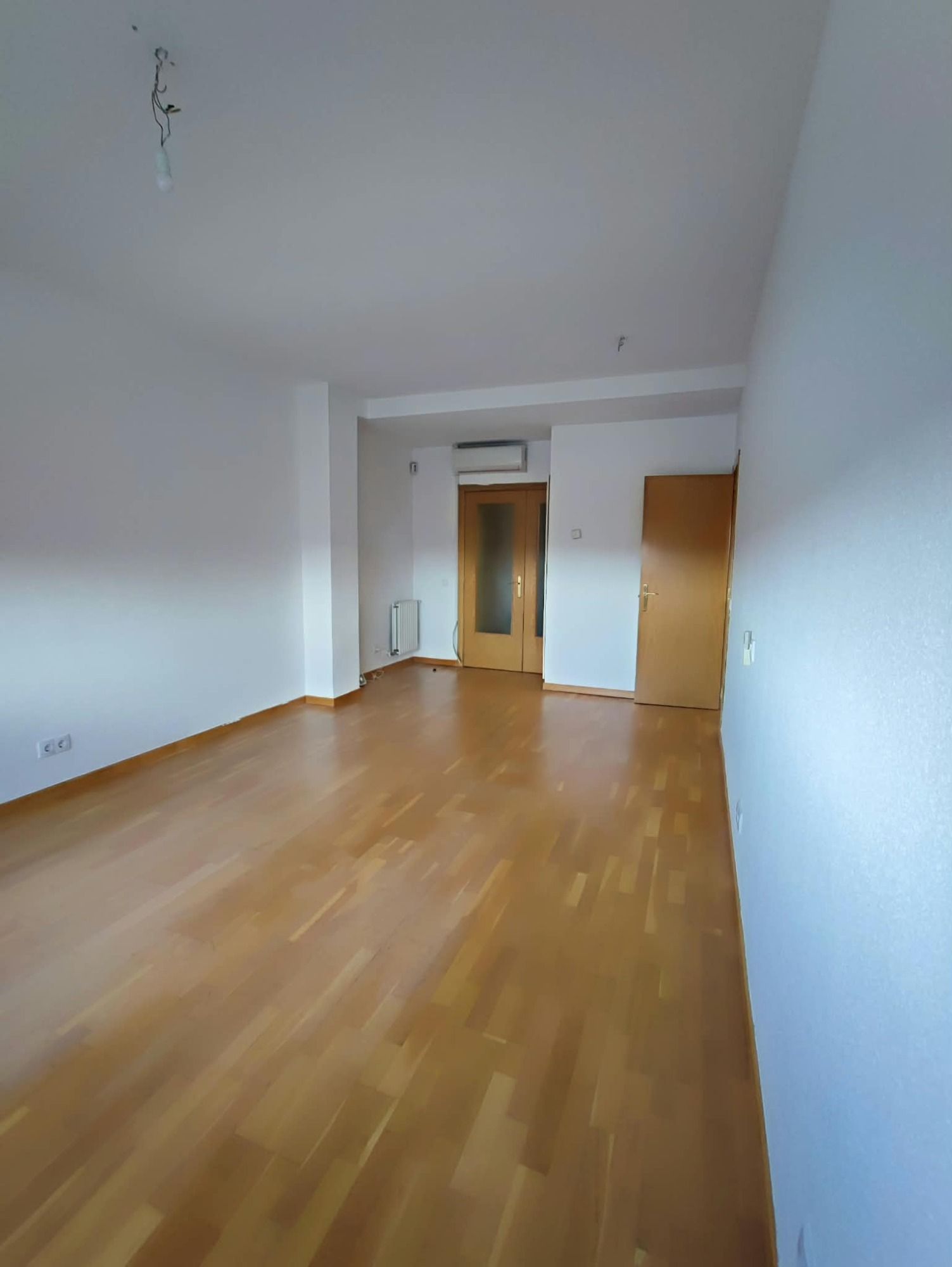 Habitación blanca vacía con suelo de madera, puertas al fondo y un radiador de pared.