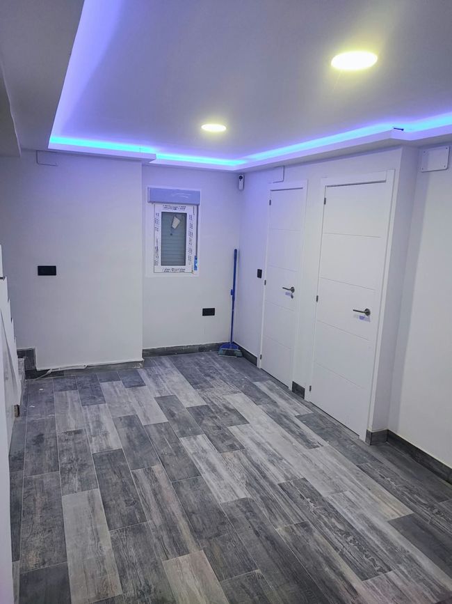 Habitación vacía con suelo gris imitación madera, paredes blancas, luces LED azules en el techo y dos puertas blancas.