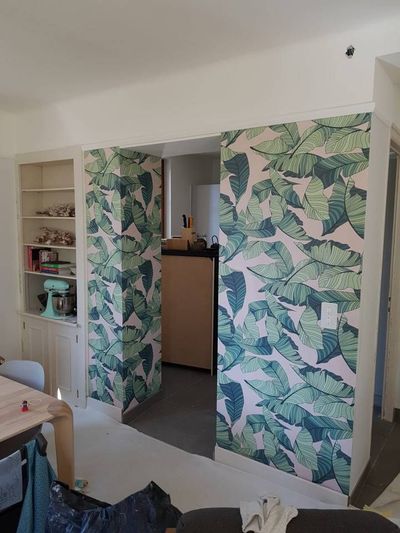 Petite pièce avec papier peint à motifs de feuillage, porte ouverte, étagères et table au premier plan.
