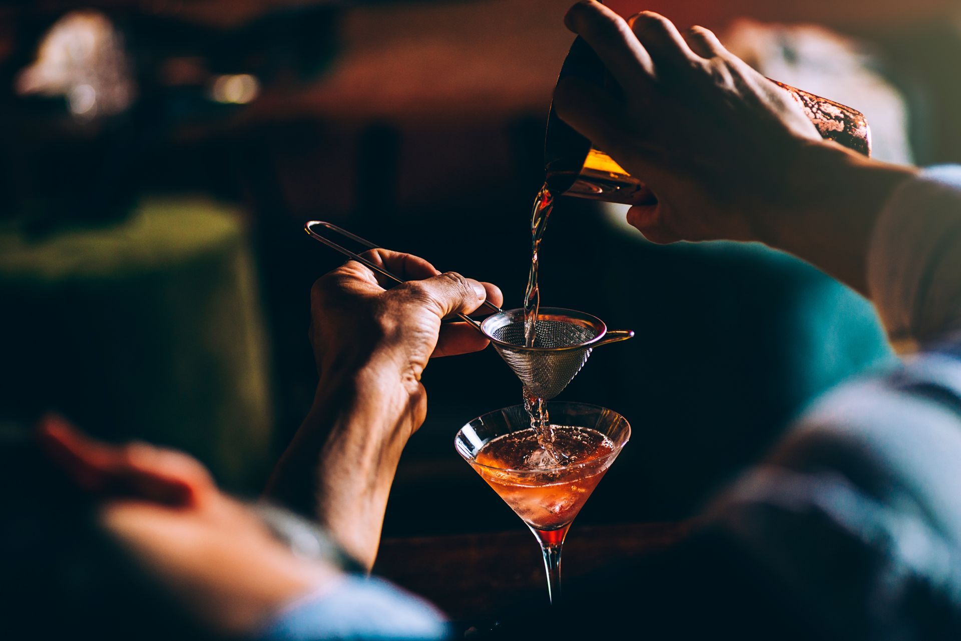 Dans un bar à l'éclairage tamisé, un barman verse un cocktail rose d'un shaker en métal dans un verre à martini.