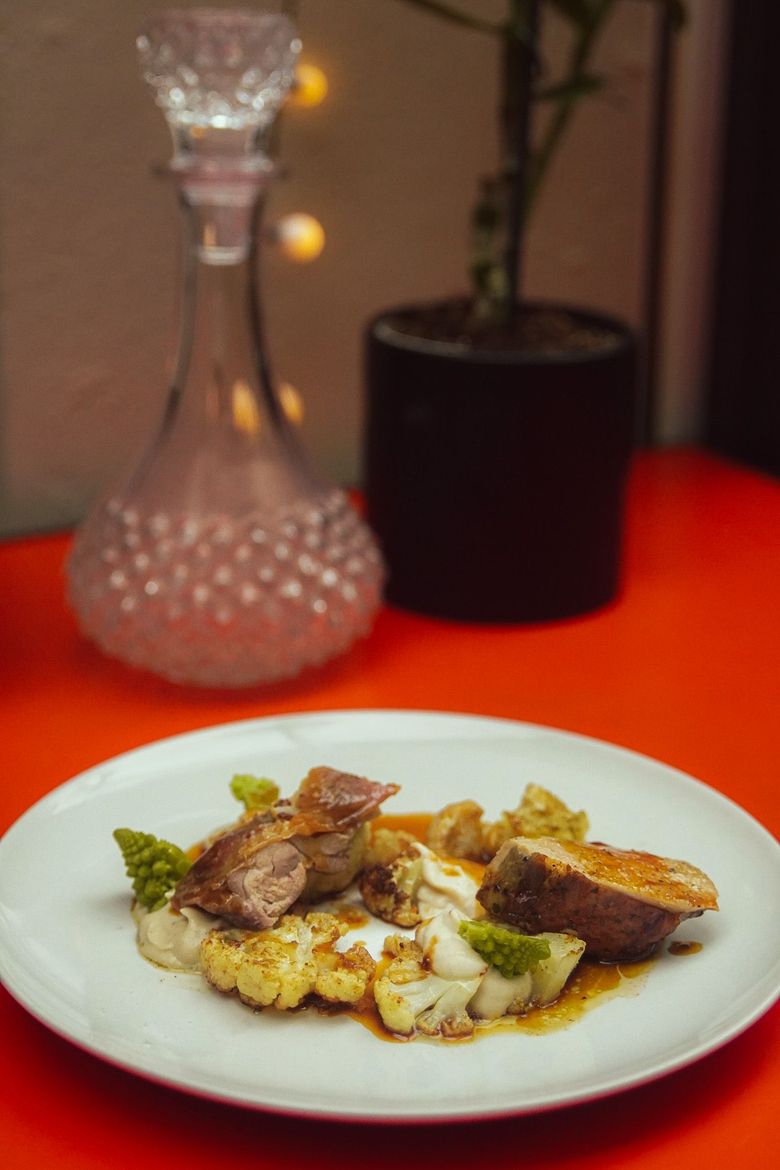 Une personne verse du vin d'une bouteille à l'étiquette orange vif dans un verre à pied tenu par une autre personne à table.