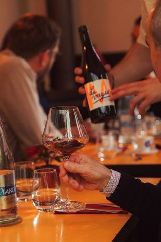 Une personne verse du vin d'une bouteille à l'étiquette orange vif dans un verre à pied tenu par une autre personne à table.