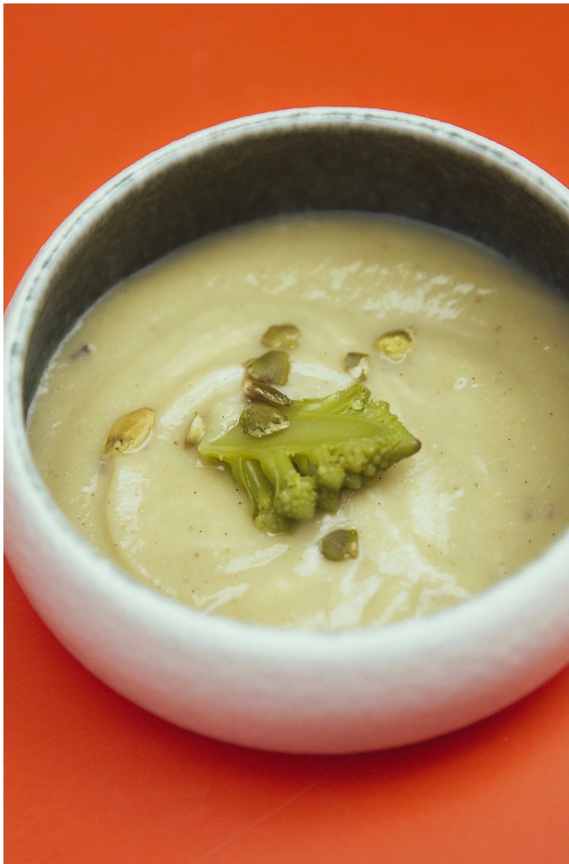 Un bol de soupe onctueuse et crémeuse, garni d'un seul morceau de brocoli et de graines, sur un fond orange vif.