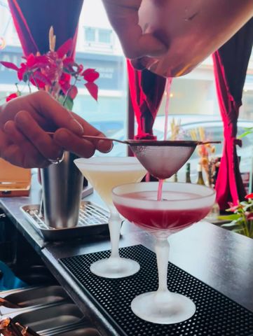 Un cocktail à la couleur sombre, surmonté d'une épaisse couche de mousse blanche, servi dans un verre à pied sur un sous-verre, avec un arrière-plan de bar flou.