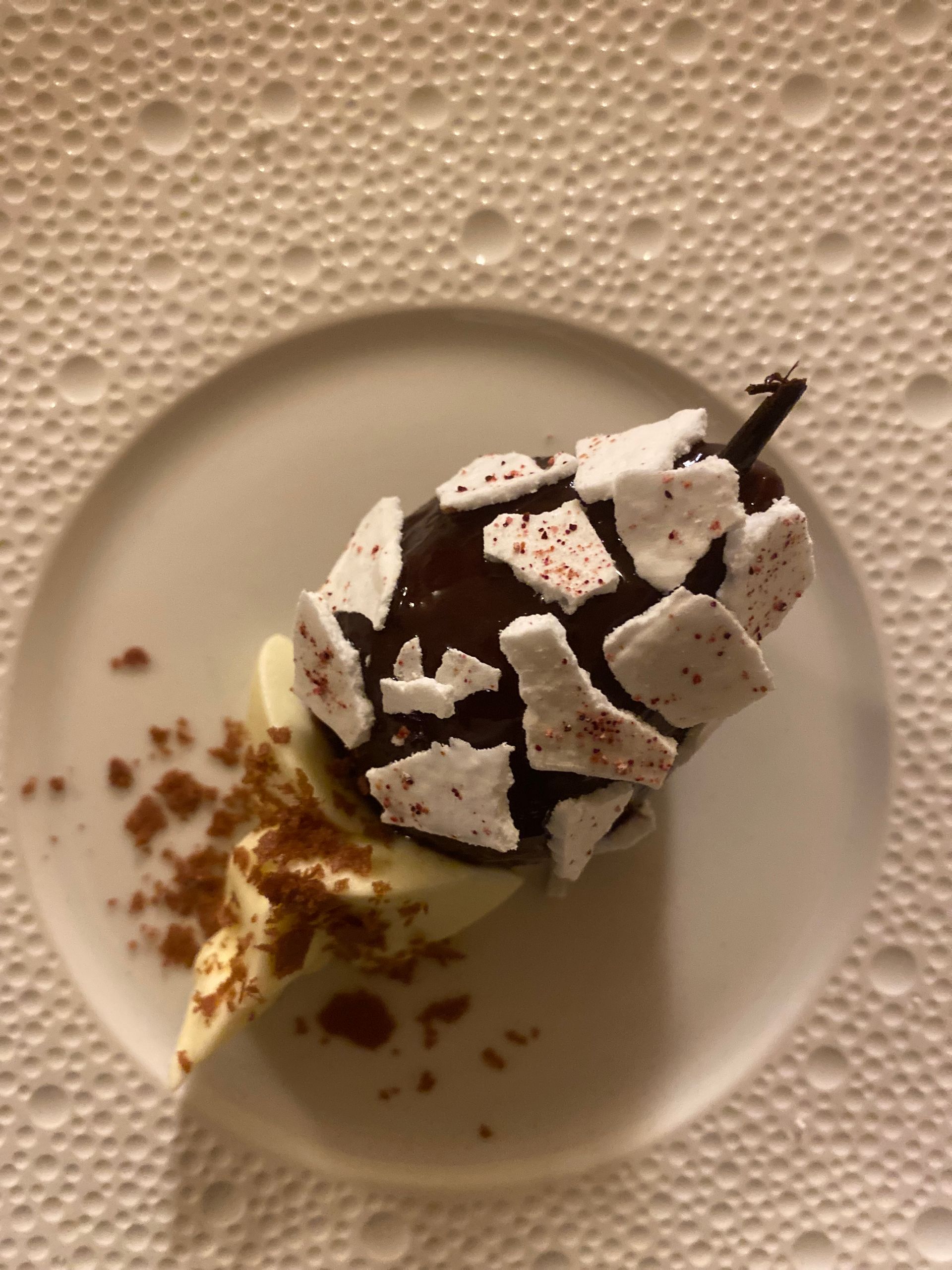 Une poire enrobée de chocolat, garnie d'éclats de meringue blanche et de poudre de cacao, servie sur un lit de crème dans une assiette texturée.