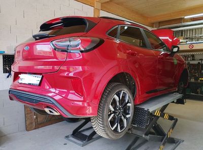 SUV rouge sur un pont élévateur dans un garage, vue arrière gauche.