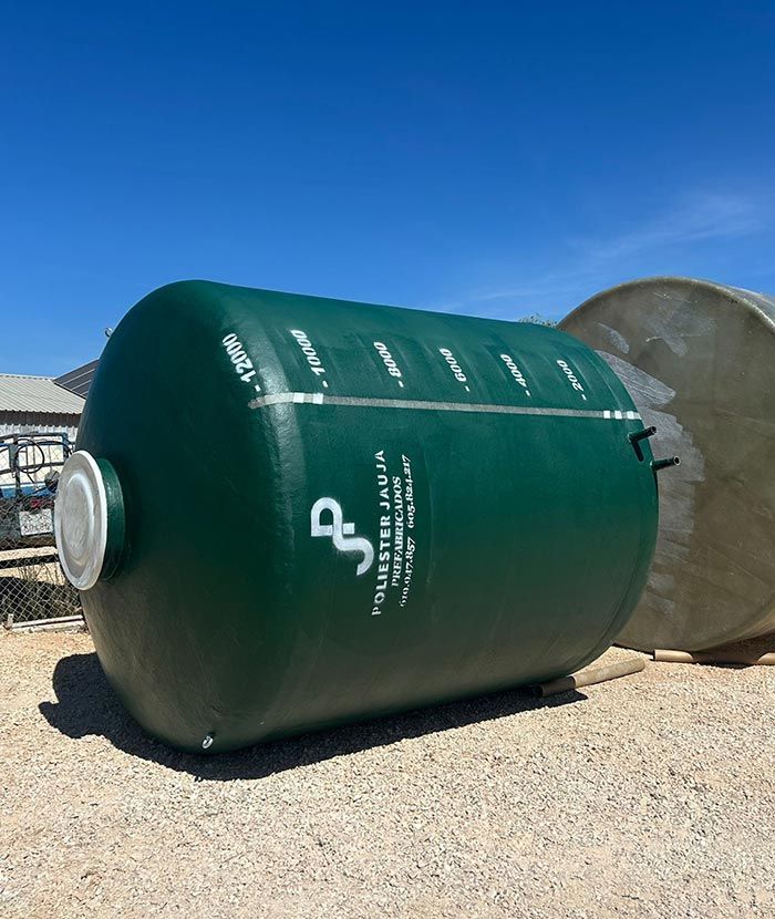Tanque cilíndrico verde sobre un lote de grava, con tapa y marcas blancas, a plena luz del sol.