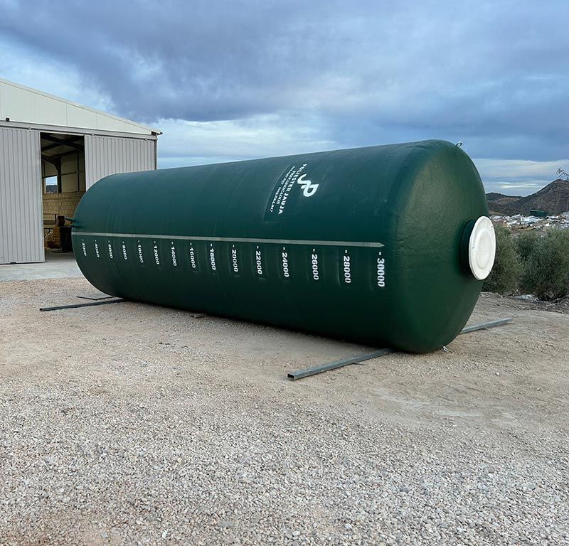 Tanque de almacenamiento de agua cilíndrico verde sobre una superficie de grava, afuera de un edificio.