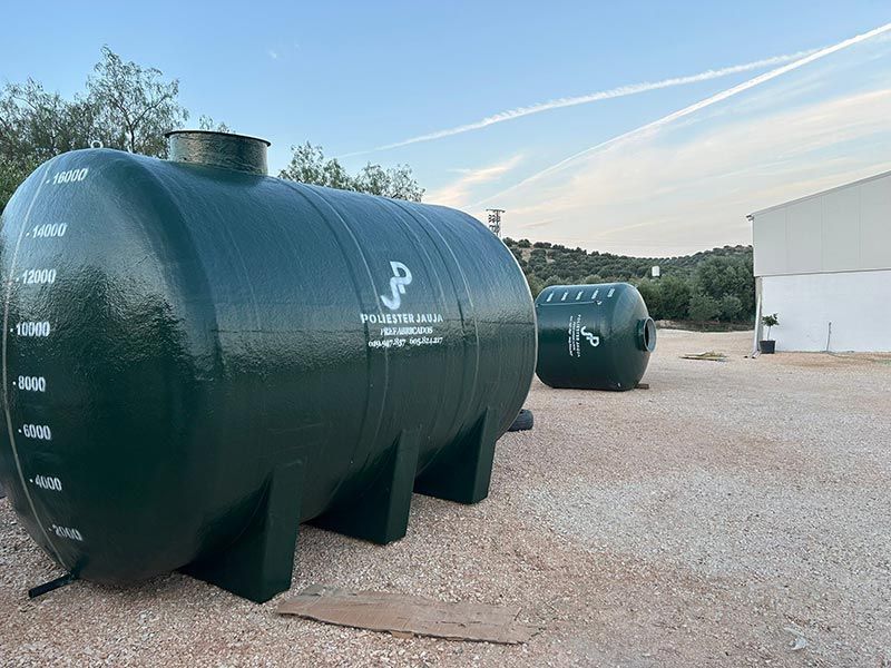 Dos grandes tanques de agua cilíndricos verdes con etiquetas, sobre grava. Cielo azul.