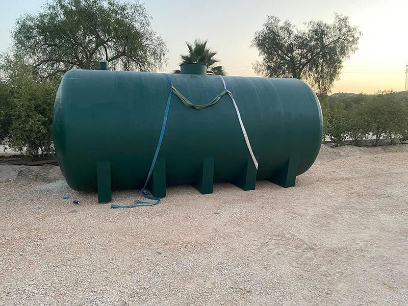 Tanque de almacenamiento cilíndrico verde apoyado sobre soportes de hormigón, al aire libre sobre grava, con árboles de fondo.