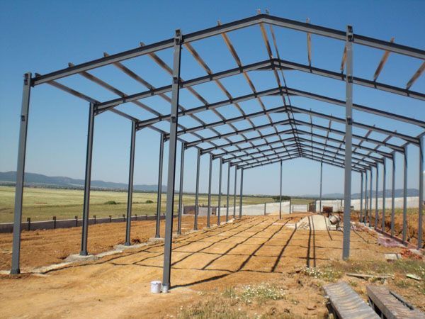 Estructura de acero de un granero en construcción en un terreno baldío, con campos al fondo.