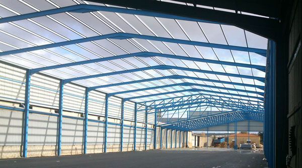 Gran almacén industrial vacío con vigas de acero azul en el techo y luz solar que se filtra a través de paneles translúcidos.