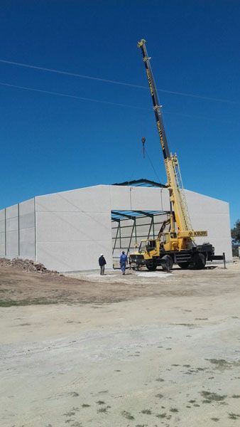 Una grúa eleva una estructura de acero junto a un gran edificio industrial blanco bajo un cielo azul despejado.