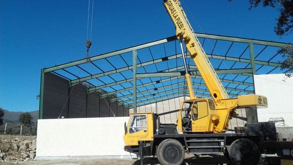 Una grúa amarilla levanta vigas de acero en una obra de construcción junto a un edificio con estructura metálica y paredes blancas.