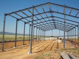 Estructura de acero de un almacén en construcción en un terreno baldío, con campos abiertos al fondo.
