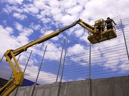 Trabajadores en una plataforma elevadora amarilla inspeccionan una valla contra un cielo azul nublado.
