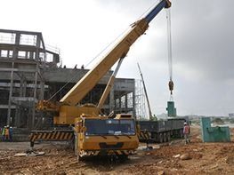 Una grúa amarilla levanta un contenedor en una obra en construcción embarrada junto a la estructura de un edificio de acero.