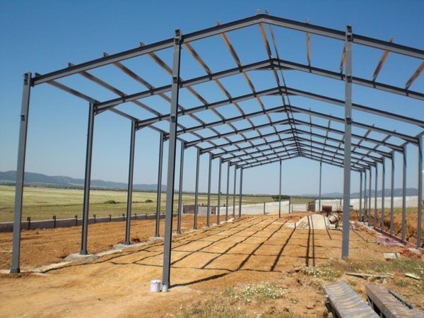 Estructura de acero de un granero en construcción en un terreno baldío, con campos al fondo.
