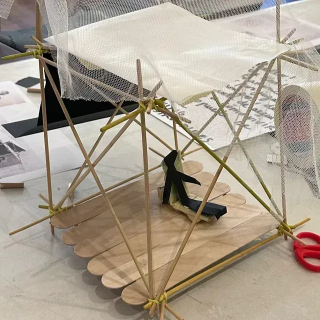 Modelo de una balsa hecha con palitos de helado con un toldo de malla y un asiento negro, en el interior.