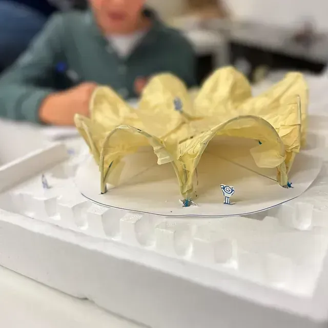 Maqueta arquitectónica de una estructura arqueada de color amarillo con pequeñas figuras sobre una base circular blanca, con una persona al fondo.