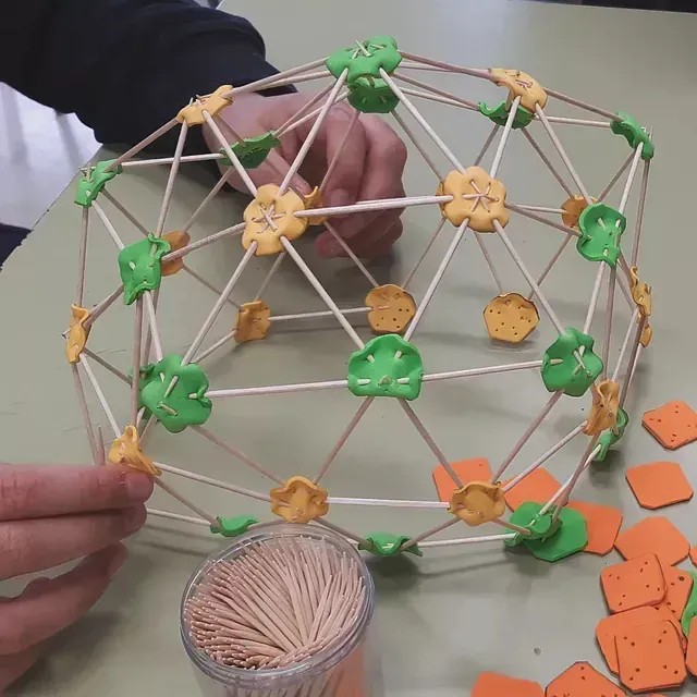 Persona ensamblando una cúpula geodésica con palillos de dientes y arcilla de colores en una mesa.