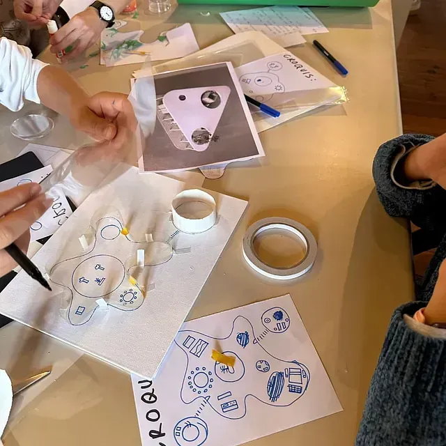 Niños en una mesa dibujando y trabajando en un proyecto de manualidades; se ven planos, herramientas y materiales.