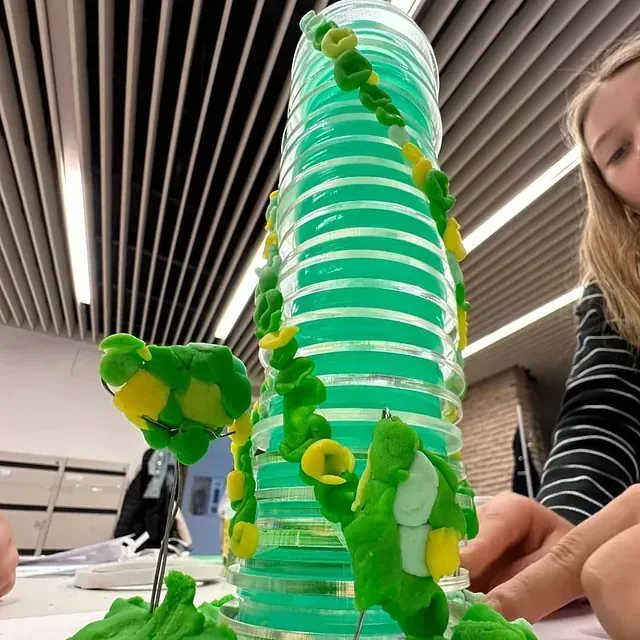Una escultura de arcilla verde y amarilla de una torre con enredaderas, sobre una mesa; una persona es visible a la derecha.