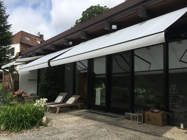 Weiße Markisen spenden Schatten über einem Gebäude mit großen Fenstern. Zwei Liegestühle stehen auf einer Terrasse.