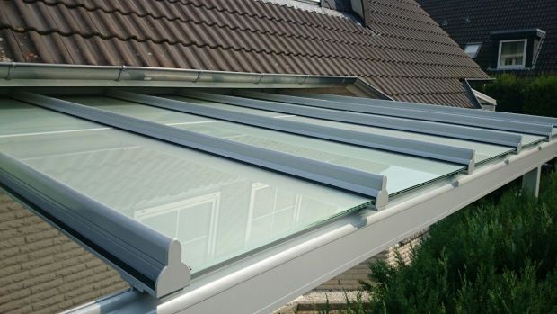 Glasdachmarkise mit weißem Rahmen und braunem Ziegeldach im Hintergrund.