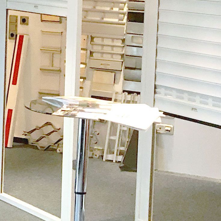 Ein silberner Tisch mit Papieren steht vor einer Glaswand. Regale und ein teilweise geöffneter Fensterladen sind zu sehen.