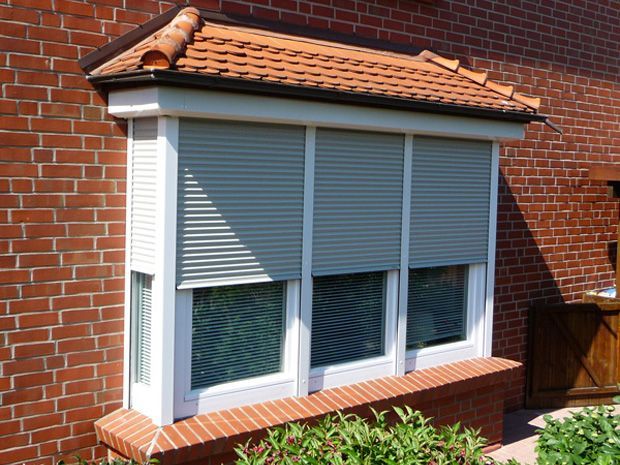 Ein Erkerfenster mit drei Scheiben, grauen Rollläden und einer Backsteinfassade.