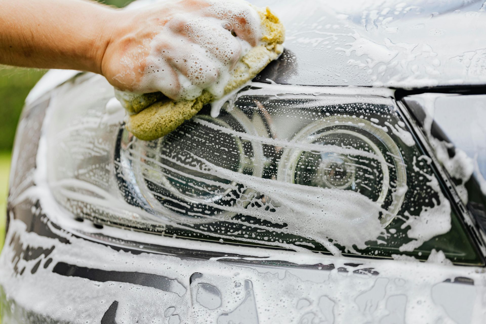 A hand uses a soapy yellow sponge to wash the headlight of a gray car.