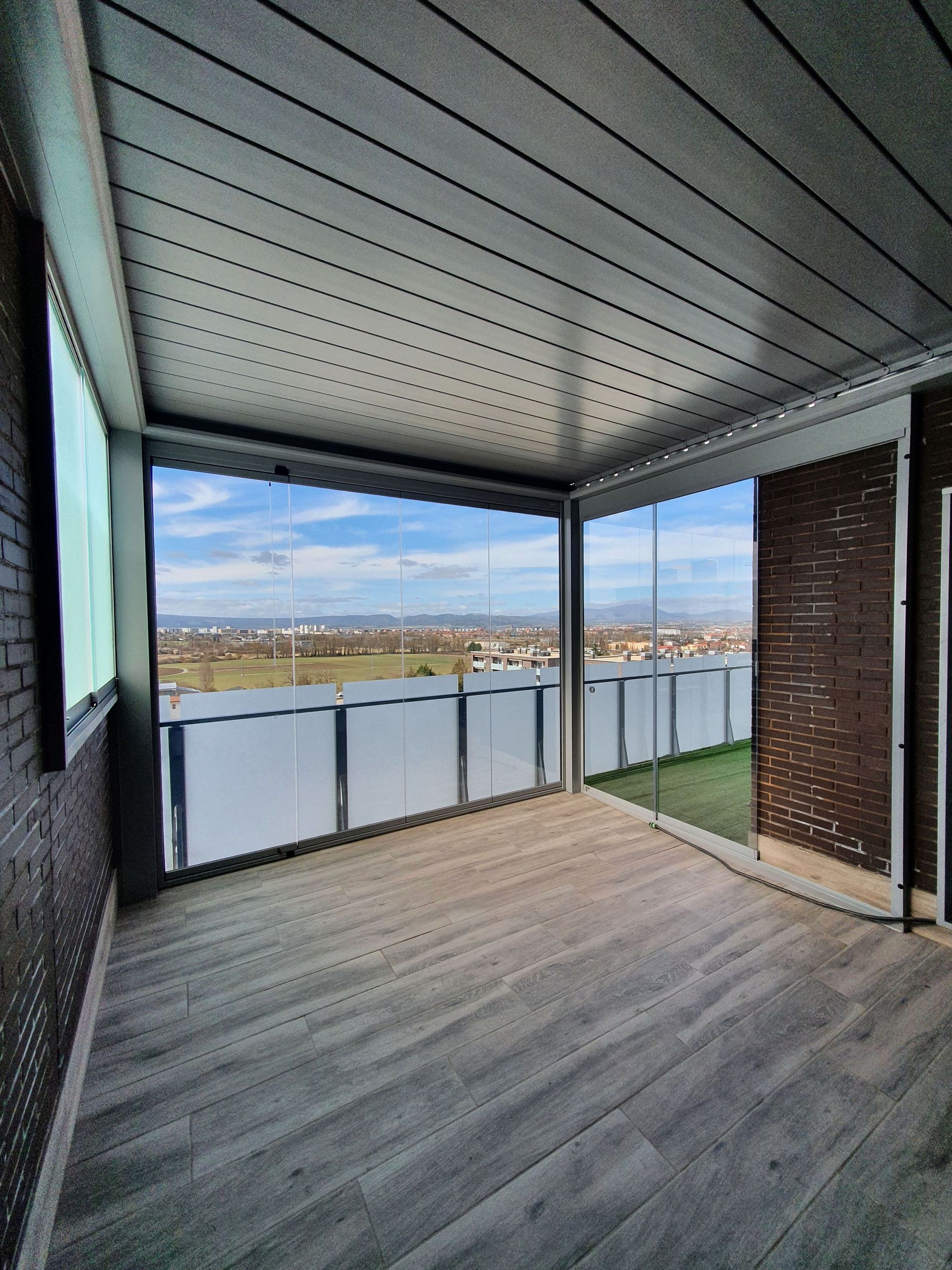 Un balcón cerrado con suelo de imitación madera, paredes de ladrillo y barandillas de cristal con vistas a un paisaje abierto.