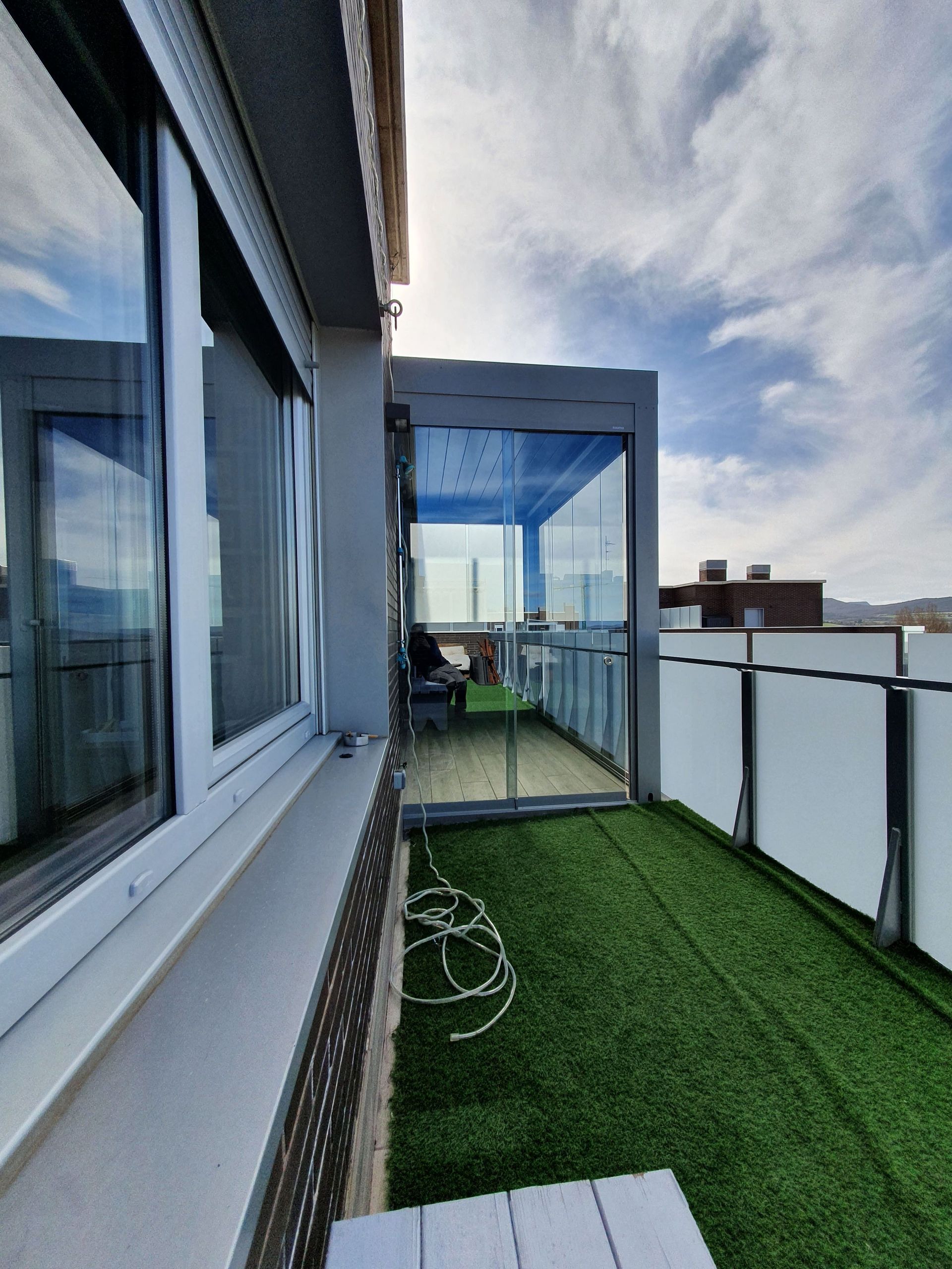 Un balcón con césped artificial, una zona de estar acristalada y una barandilla blanca contra un cielo azul con nubes ligeras.
