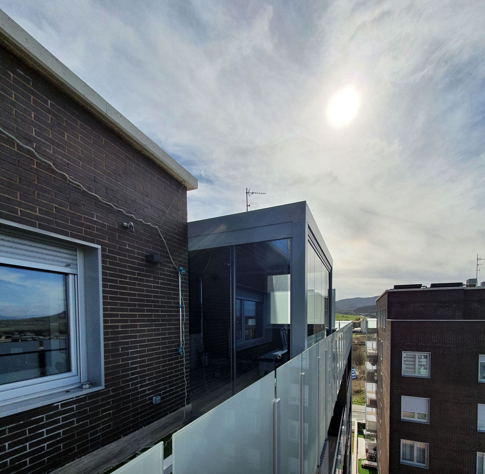 Vista de un día soleado desde un balcón con vistas a una moderna ampliación de cristal en un edificio de ladrillo junto a otro bloque de apartamentos.