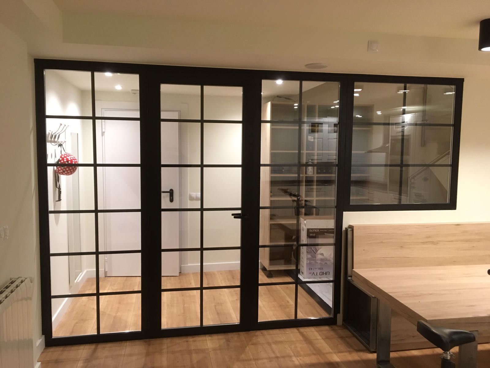 Mampara de cristal con marco negro y puerta central abatible que separa una habitación con suelo de madera de un espacio de trabajo contiguo.