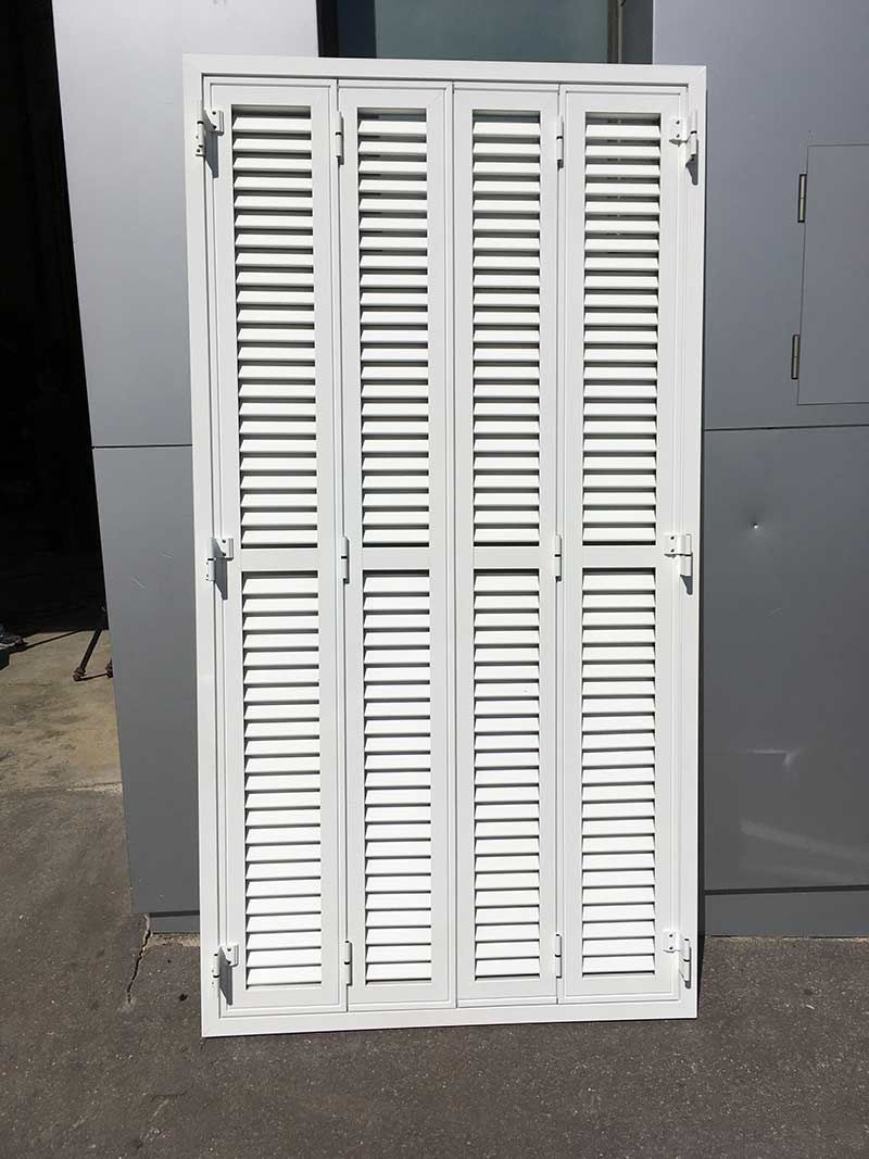 Una persiana plegable blanca de cuatro paneles, con lamas, se yergue contra una pared gris.