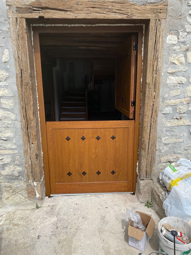 Una puerta de madera de estilo holandés con calados decorativos en forma de diamante, integrada en una pared de piedra, conduce a una oscura escalera interior.