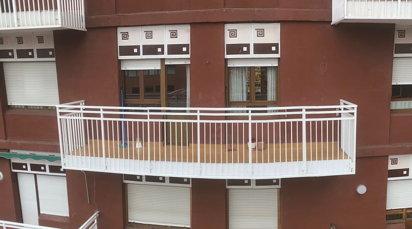 Fachada de un edificio de color marrón rojizo con un gran balcón con barandilla blanca y varias persianas enrollables blancas cerradas.