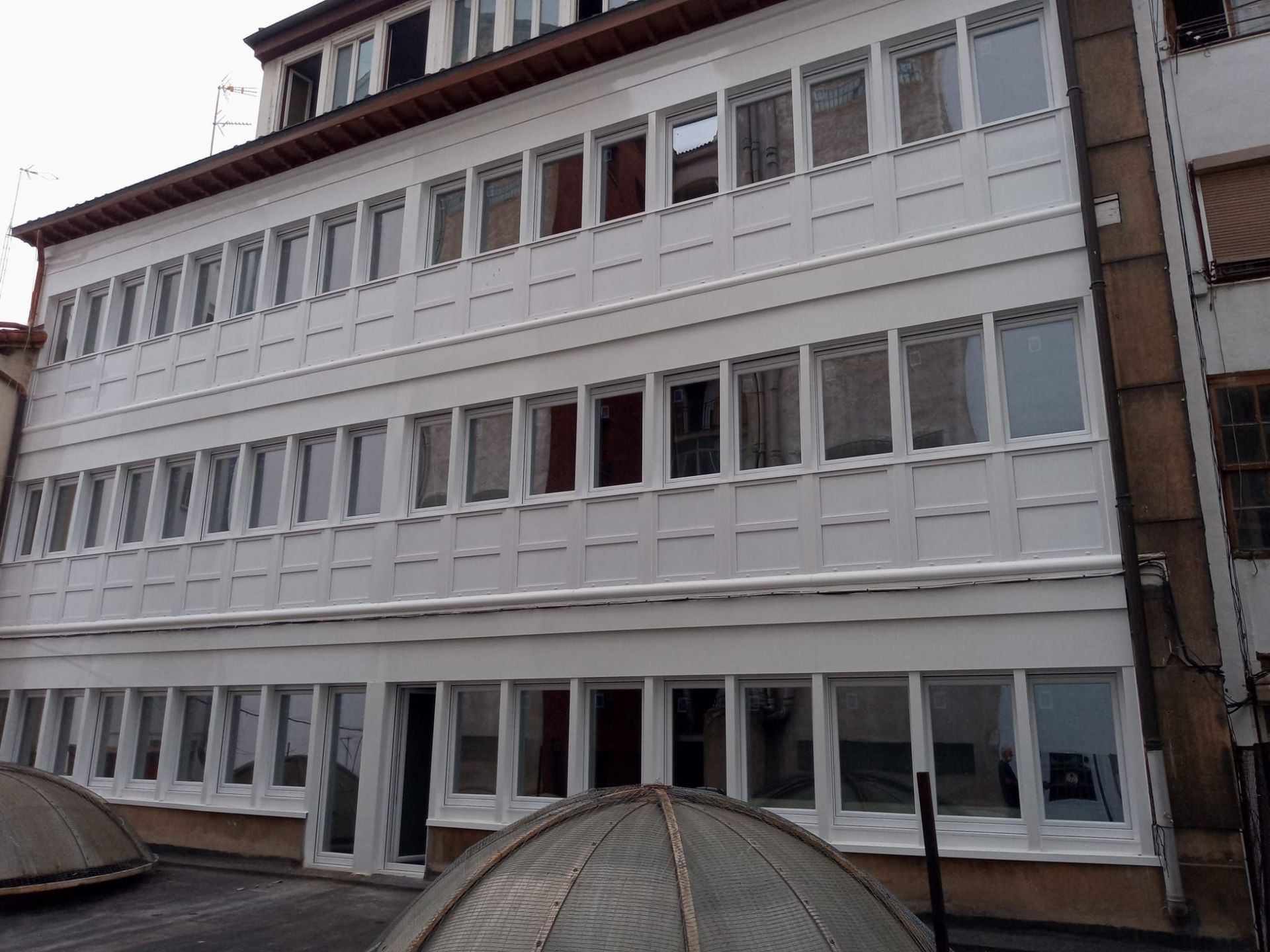 Edificio de fachada blanca con tres niveles de ventanas continuas de múltiples paneles y una cúpula de cristal en primer plano.