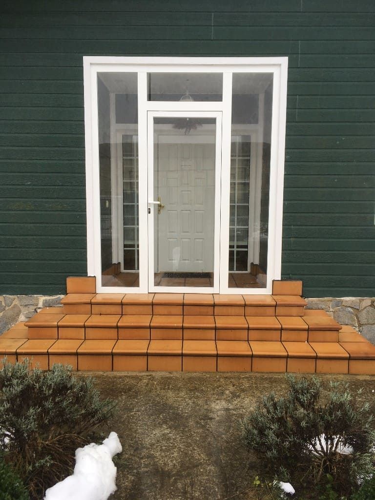 Una puerta con paneles de vidrio blanco enclavada en la fachada verde oscuro de una casa, con tres escalones de baldosas que conducen a la entrada.