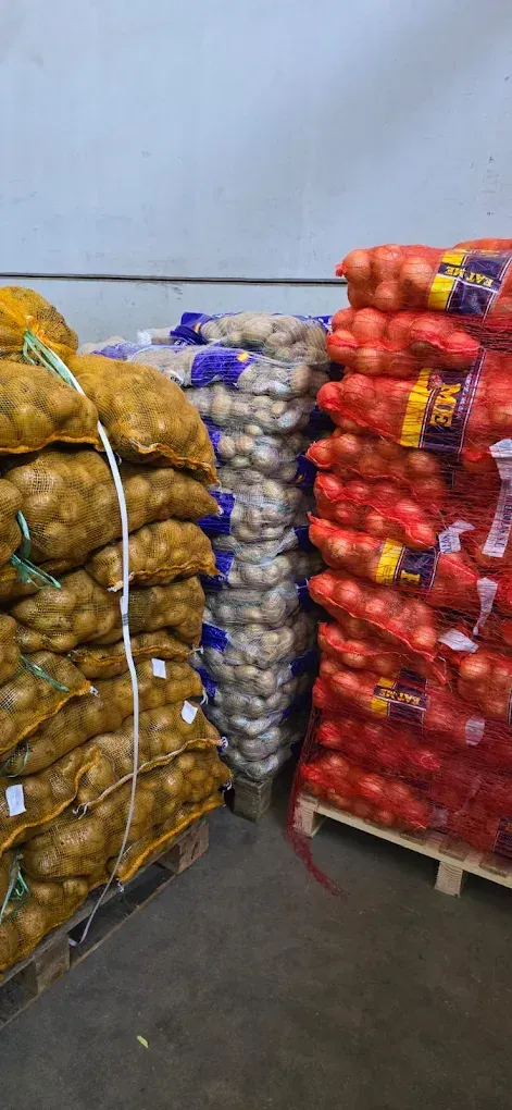 Pilas de cebollas en bolsas de malla de color rojo, blanco y amarillo se encuentran sobre paletas en un almacén.