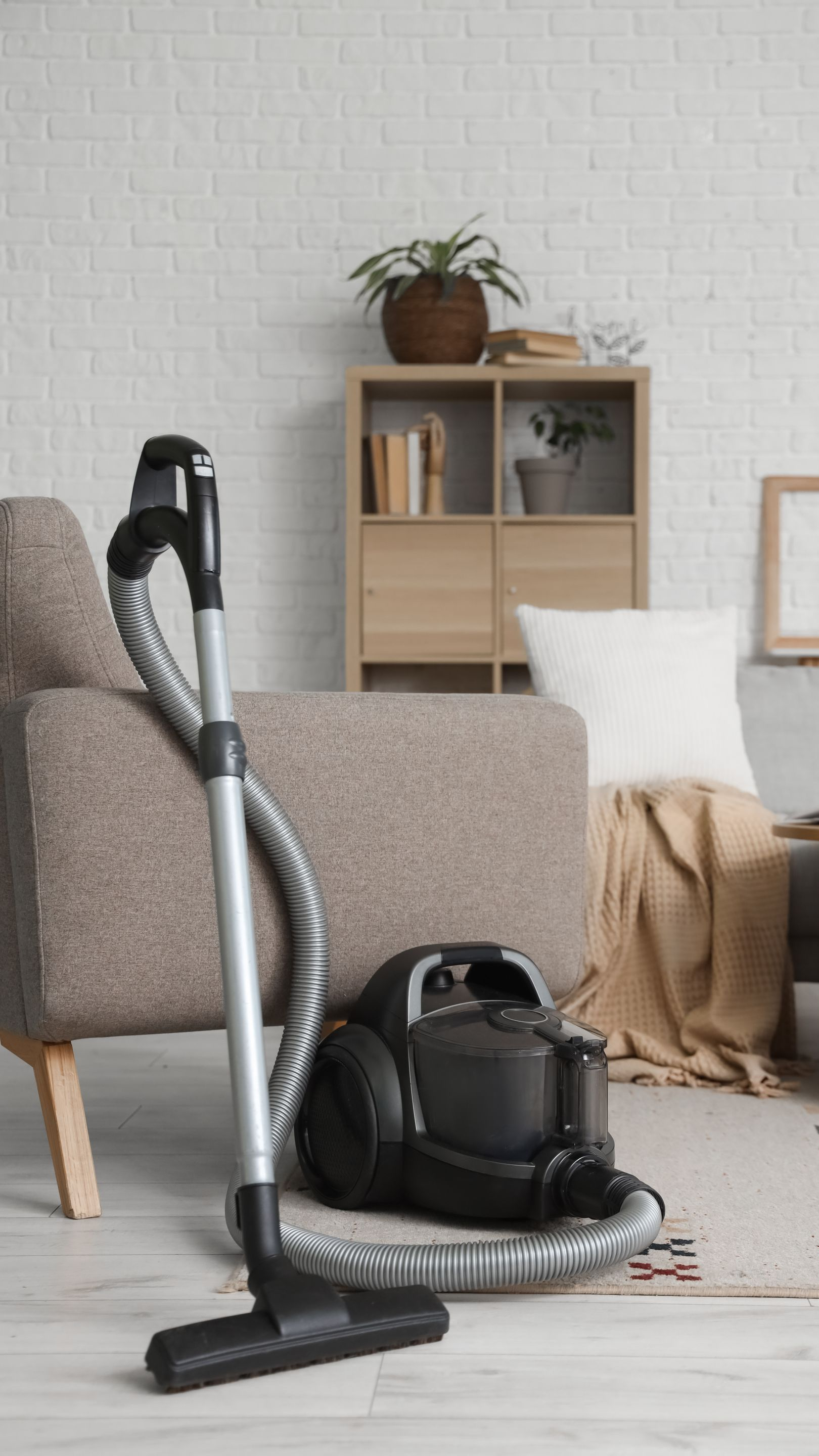Un aspirateur noir à côté d'un canapé beige dans une pièce avec un tapis clair et une petite bibliothèque en bois.