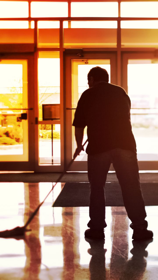 Une personne lave un sol brillant près de portes vitrées ; la lumière du soleil inonde la pièce, illuminant la scène.