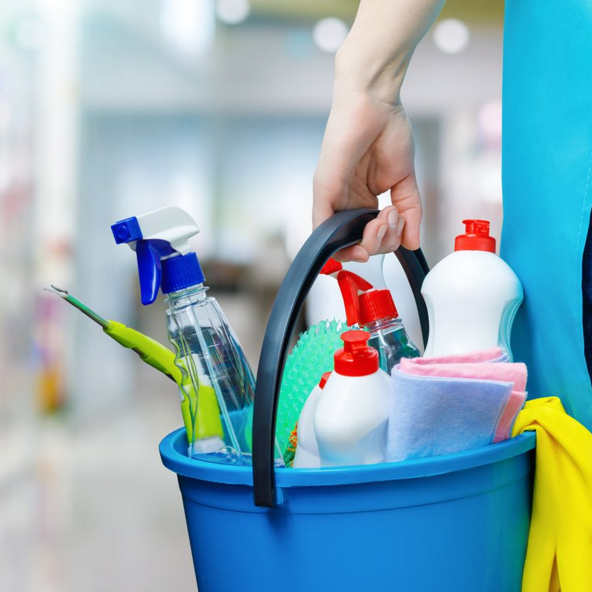 Main tenant un seau bleu rempli de produits de nettoyage, notamment des flacons pulvérisateurs et des éponges.