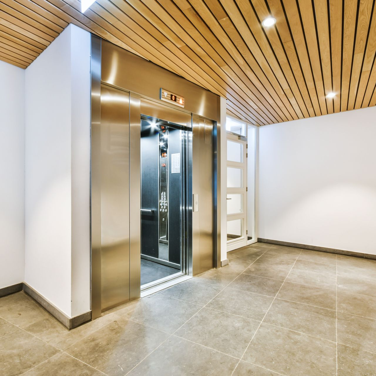 L'ascenseur, dont les portes sont en acier inoxydable, s'ouvre sur un couloir au plafond en bois clair et au sol carrelé de gris.