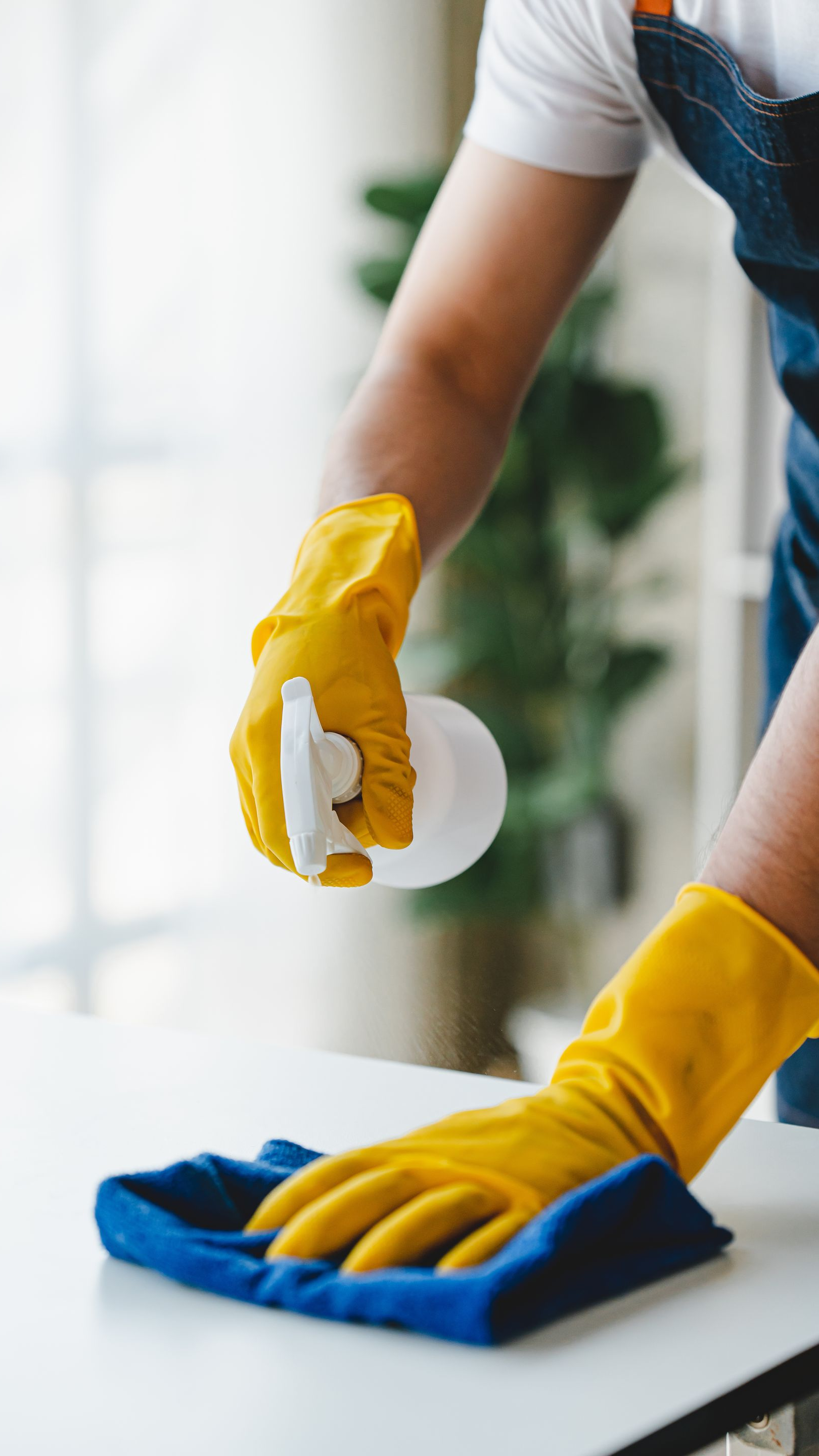 Une personne portant des gants jaunes vaporise et essuie une surface blanche avec un chiffon bleu.