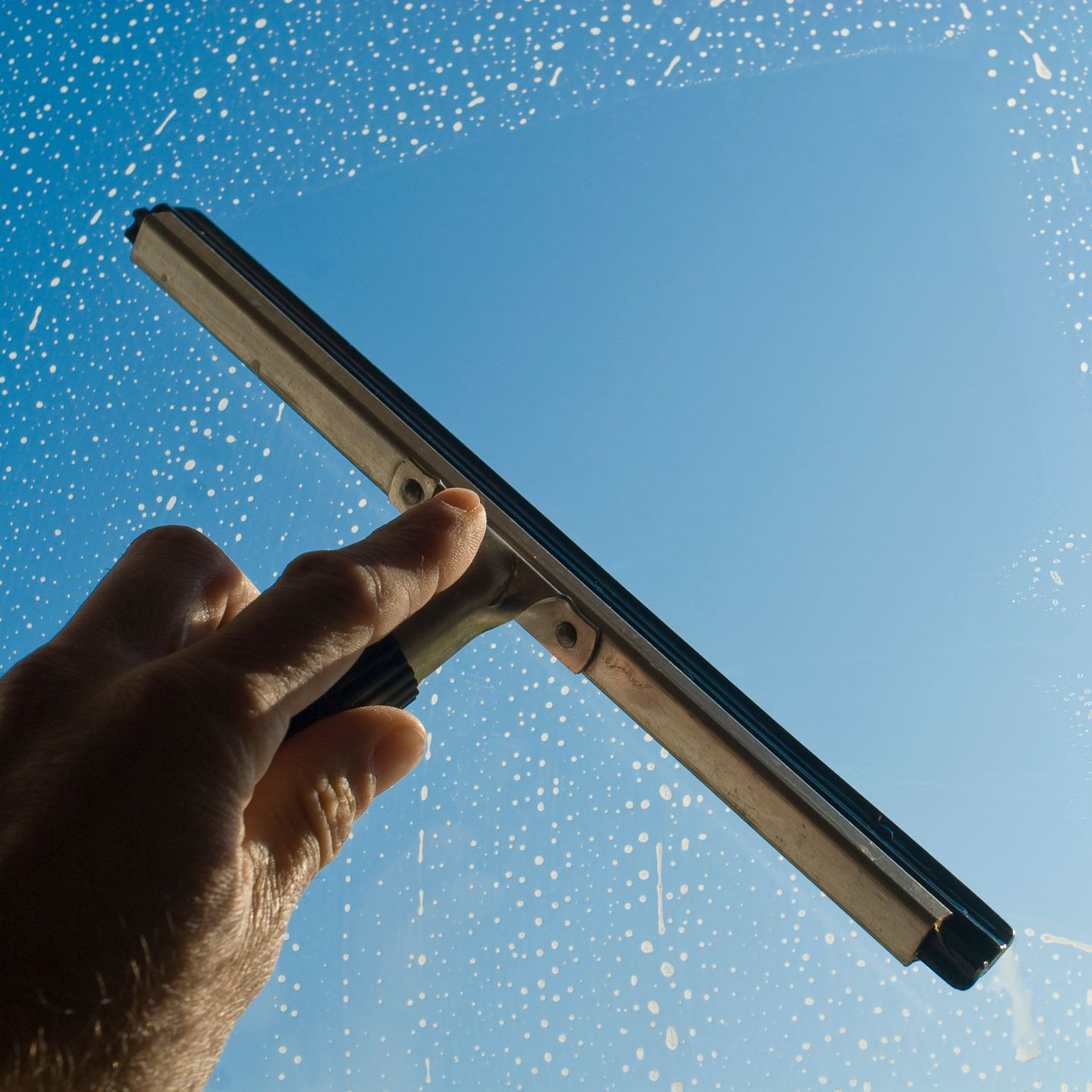 Main utilisant une raclette pour nettoyer une vitre sur fond de ciel bleu.